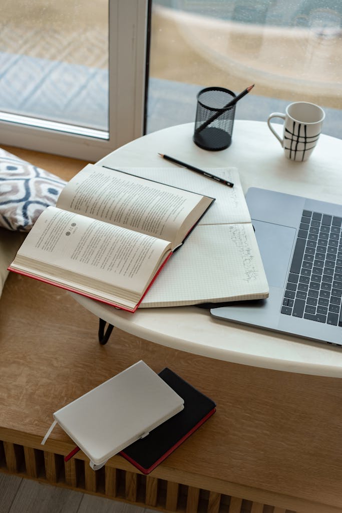 A cozy indoor study setup featuring an open book, laptop, and coffee mug by the window.